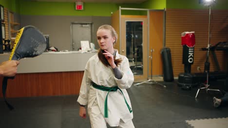 a young girl combat fighter taekwondo karate martial arts student does slow motion practice training spinning high kick into pad with black belt master inside a exercise strength gym studio