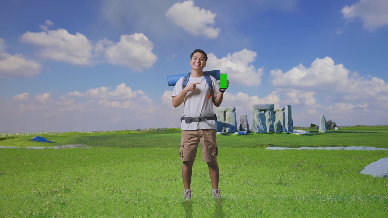 Full Body Of Asian Male Hiker With Mountaineering Backpack Smiling And Pointing To Mock Up Green Screen Smartphone While Traveling In Stonehenge