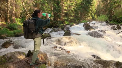 female traveler with a backpack, drinking water in nature in the forest near a mountain river.