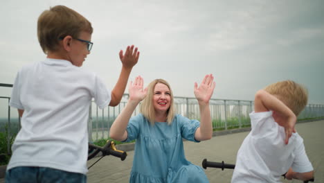 a mother and her two children share a joyful moment trying to give each other high fives, she's squatting with iron rail and greenery by the side