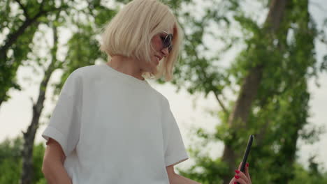 cheerful woman wearing sunglasses and white polo filming playful park scene on phone under green trees with excited expression, joyful laughter and family moment recorded in summer warmth