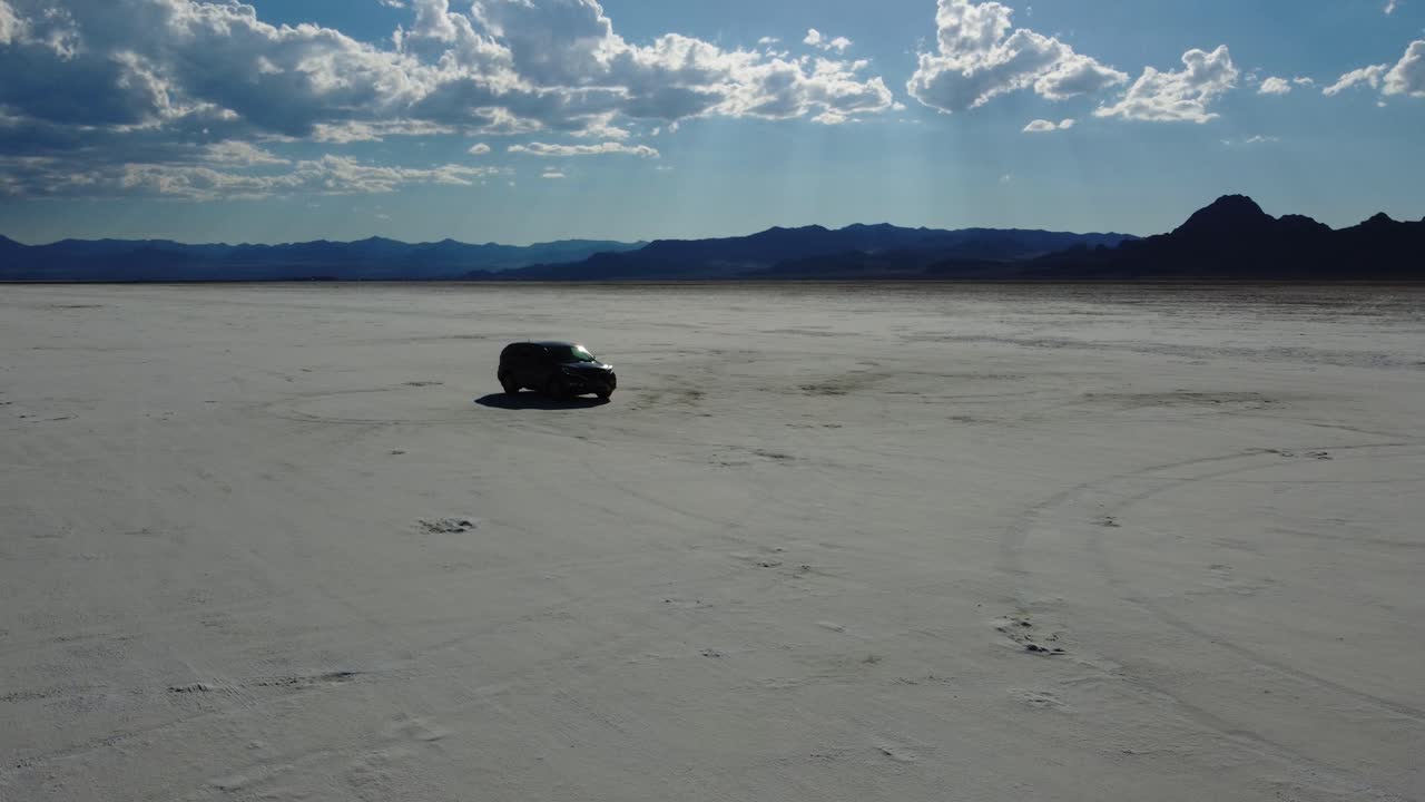 Car driving on Bonneville Salt Flats filmed from the front with 4K drone. White salt lake bed contrasts mountain range, blue sky, and sun rays peaking through the clouds. Car commercial-type shot.