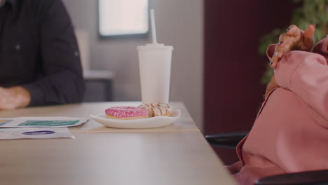 Vista-De-Cerca-De-Una-Mujer-Embarazada-Sentada-En-La-Mesa-Comiendo-Un-Donut-Y-Tocándose-El-Vientre