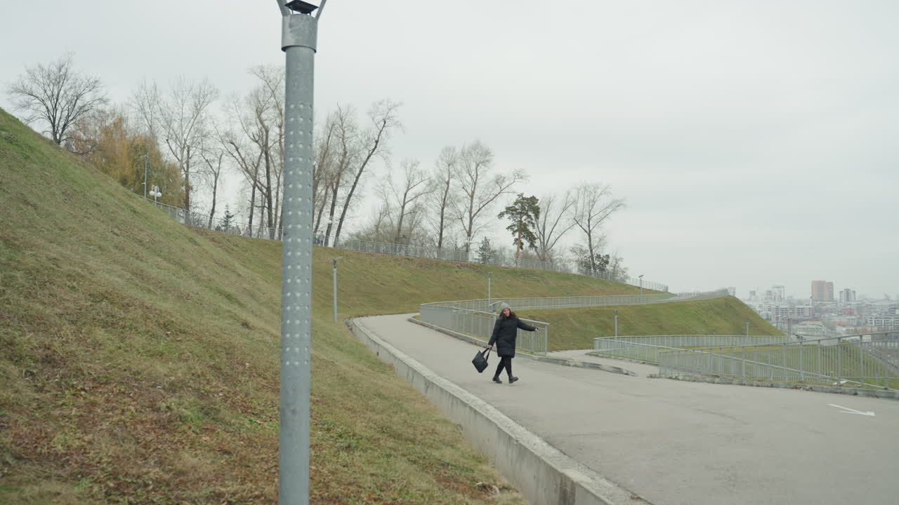 Delivery courier hurrying along empty path with package, prominent lamppost and city backdrop, brisk stride on sloped sidewalk, overcast sky and muted tones, energetic commuter vibe