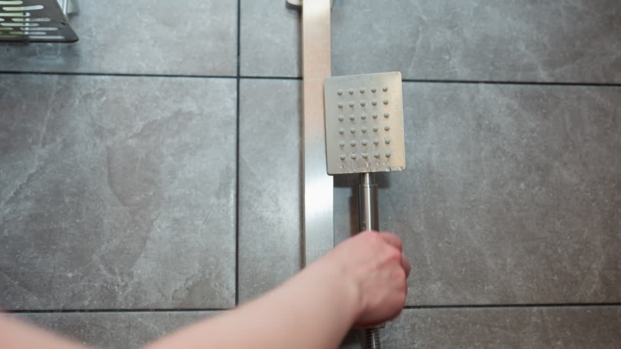 close up hand view of lady retrieving handheld shower head from wall mount before bathing capture subtle water drips and calm moment of self care in tiled wet shower environment with modern design