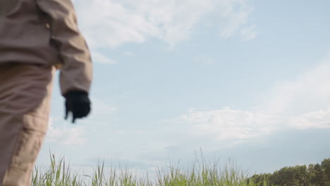 couple walking through tall green grass holding phones in hands, wearing matching khaki jackets and gloves, scanning horizon under cloudy blue sky, trees lining background
