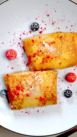 two golden nalysnyky, garnished with fresh berries, powdered sugar, and a vibrant sprinkle of red crumbs, rest on a white plate, accompanied by a glass of refreshing orange juice and a fork