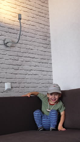 smiling caucasian toddler sitting in the corner of the sofa. lovely boy in a cap playing at home. vertical video