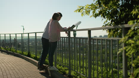 friends leaning on iron railing sharing cheerful moment as boy laughs covering face while girl points at tablet, background features distant people blurred against greenery and bright sunlight