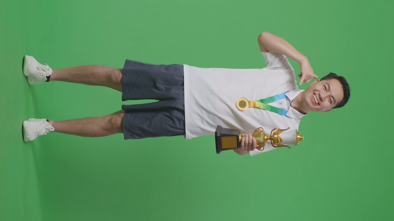 황금 메달과 트로피를 가진 아시아 남성의 몸이 가득 차 있으며, 스튜디오의 녹색 스크린 배경에서 첫 번째 우승자로서 카메라에 미소 짓는 제스처를 보여줍니다.