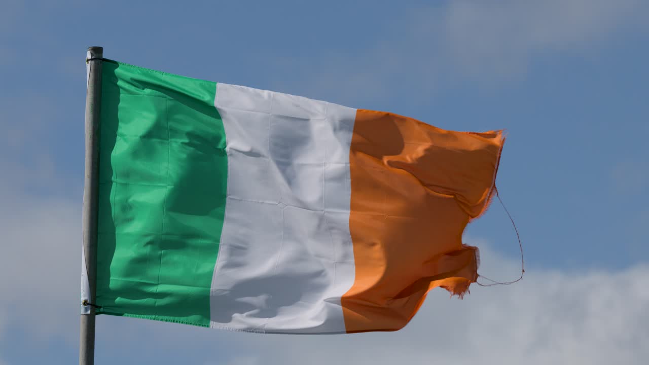 Tricolor Irish flag flutters dynamically on flagpole, bright daylight, clear sky, steady camera