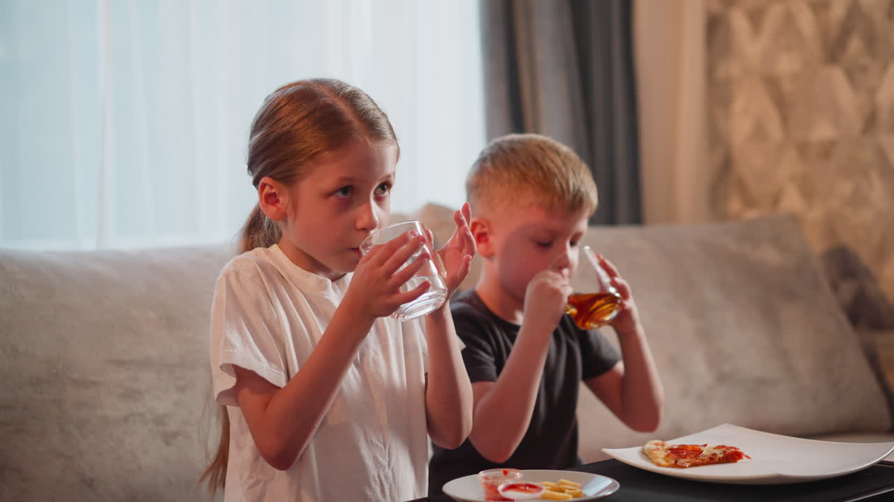 boy drinks juice girl drinks water while seated side by side on couch with plate of fries and sauce in front of girl and sliced pepperoni pizza on white plate in front of boy