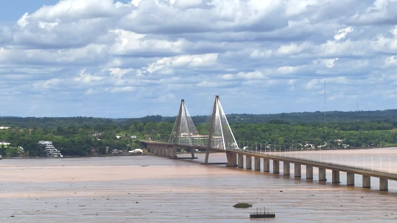 Aerial wide shot showing boat and ship on Parana River and San Roque Gonz&aacute;lez de Santa Cruz Bridge during cloudy day with forest landscape in background