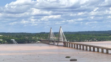 aerial wide shot showing boat and ship on parana river and san roque gonzález de santa cruz bridge during cloudy day with forest landscape in background