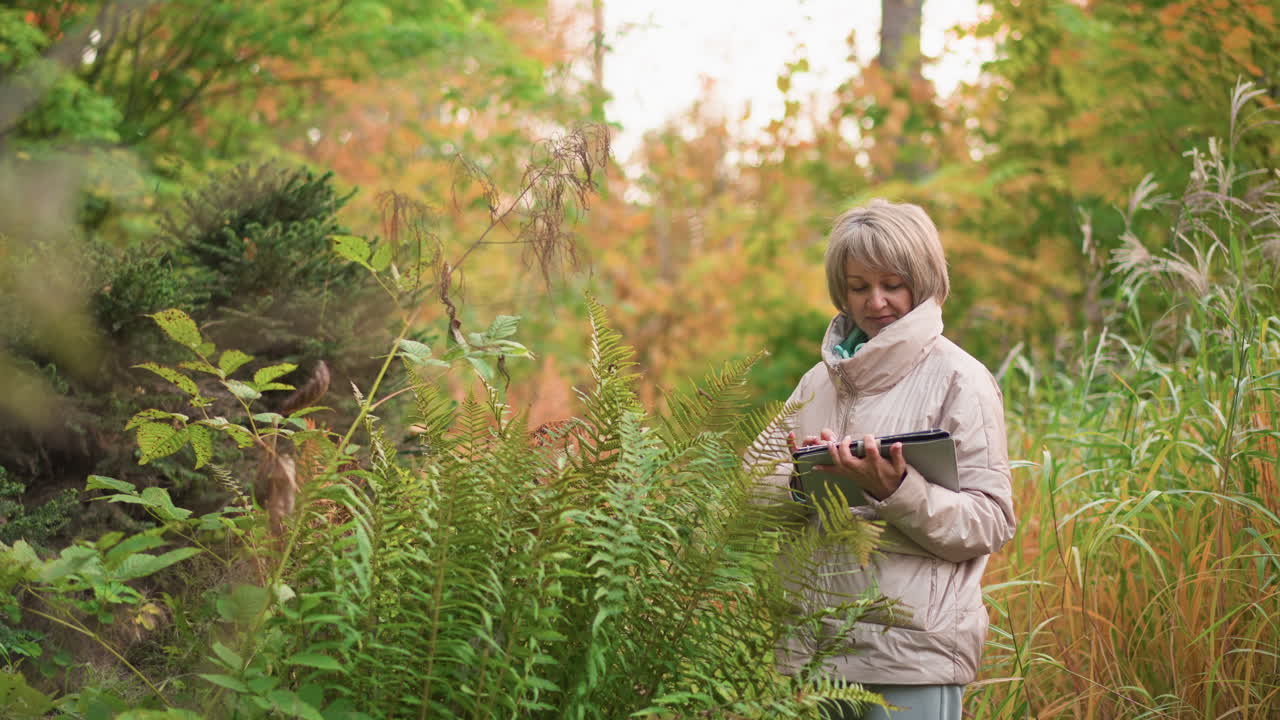 scientist in beige coat holding clipboard and writing observations while standing in thick foliage with vibrant green and orange plants, capturing peaceful moment of nature study during autumn season