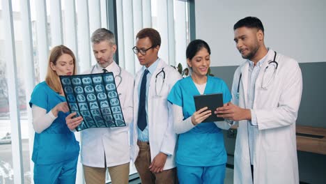 Retrato-De-Un-Equipo-Médico-De-Razas-Mixtas-Parado-En-El-Hospital-Hablando-Sobre-El-Tratamiento-De-La-Enfermedad-Del-Coronavirus,-Colegas-Masculinos-Y-Femeninos-Discuten-El-Análisis-Del-Paciente-Y-Escriben-En-Tableta,-Concepto-De-Salud