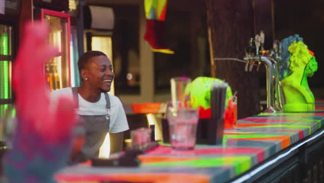 happy barman communicates with guests at bar counter. african american barkeeper shows denial and smiles in pub. joyful bartender entertains at party