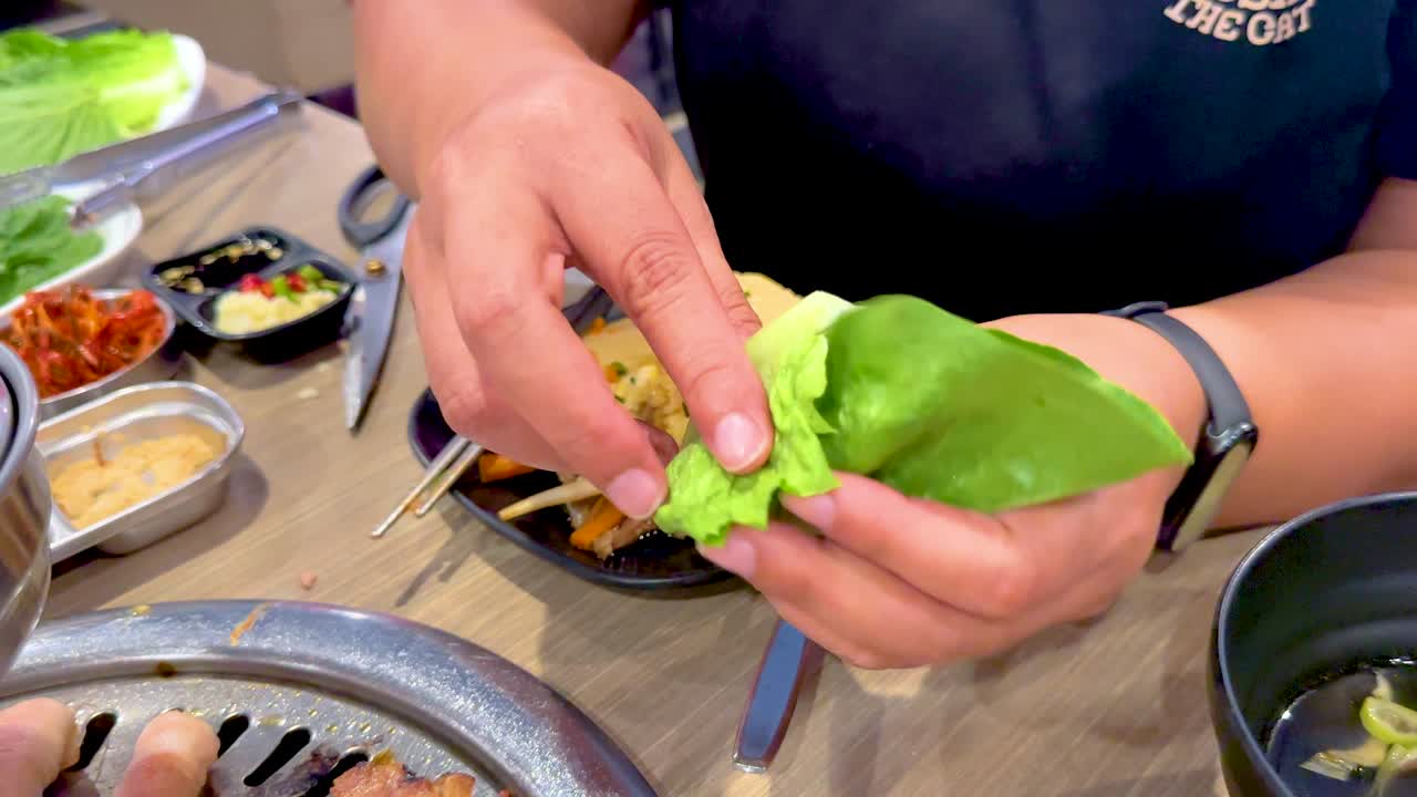 Hands assembling Korean BBQ wraps with lettuce and grilled meat in a vibrant Bangkok setting. Bright lighting enhances the culinary process