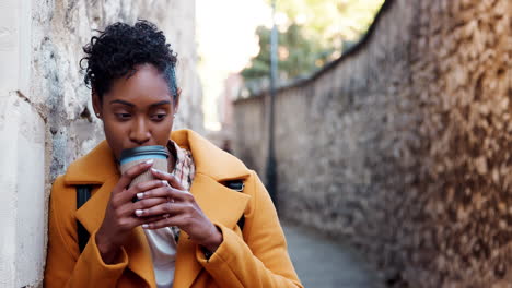 Millennial-schwarze-Frau-In-Gelbem-Mantel-Lehnt-An-Einer-Steinmauer-In-Einer-Gasse-Und-Trinkt-Einen-Kaffee-Zum-Mitnehmen,-Fokus-Auf-Den-Vordergrund