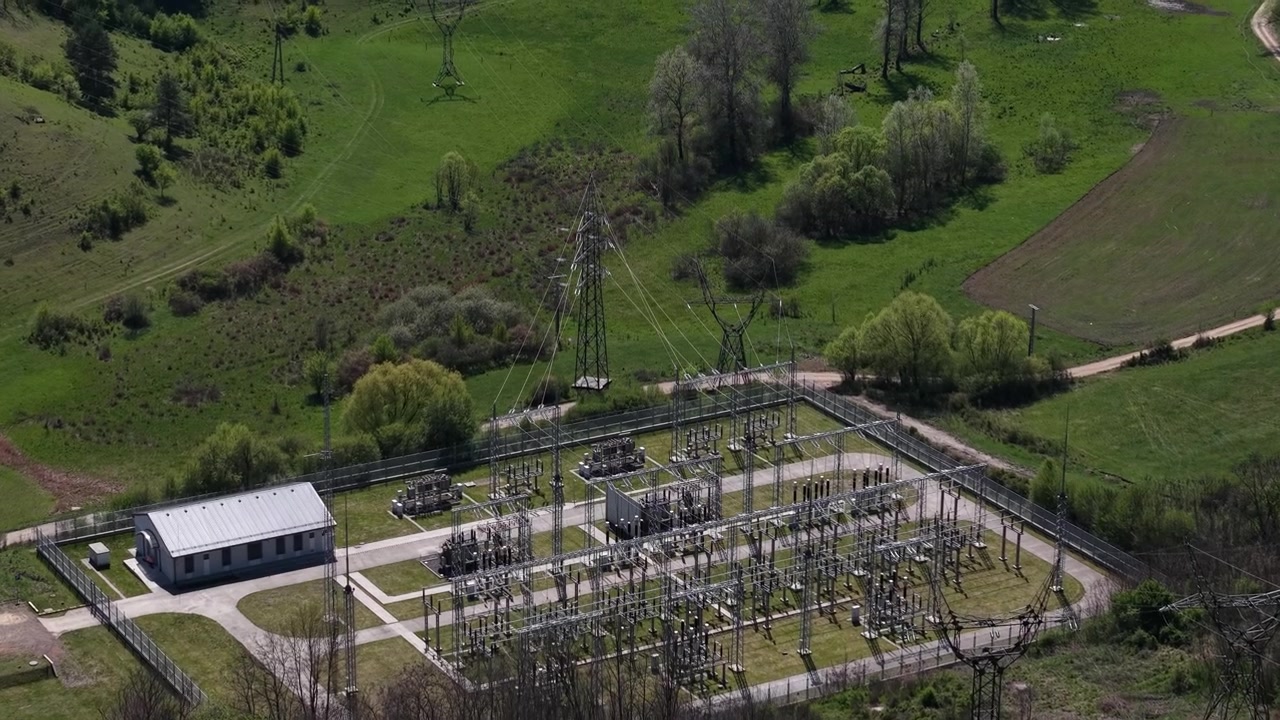 An aerial view shows an electrical substation and high-voltage power lines situated within a lush, green rural landscape.