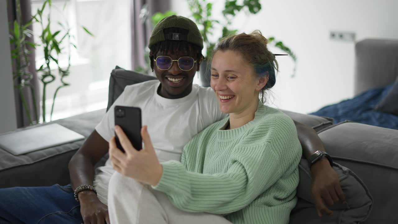 Coppia birazziale felice seduta sul divano, in videochiamata sullo smartphone.