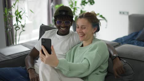 feliz pareja biracial sentada en el sofá, y teniendo una videollamada en el teléfono inteligente