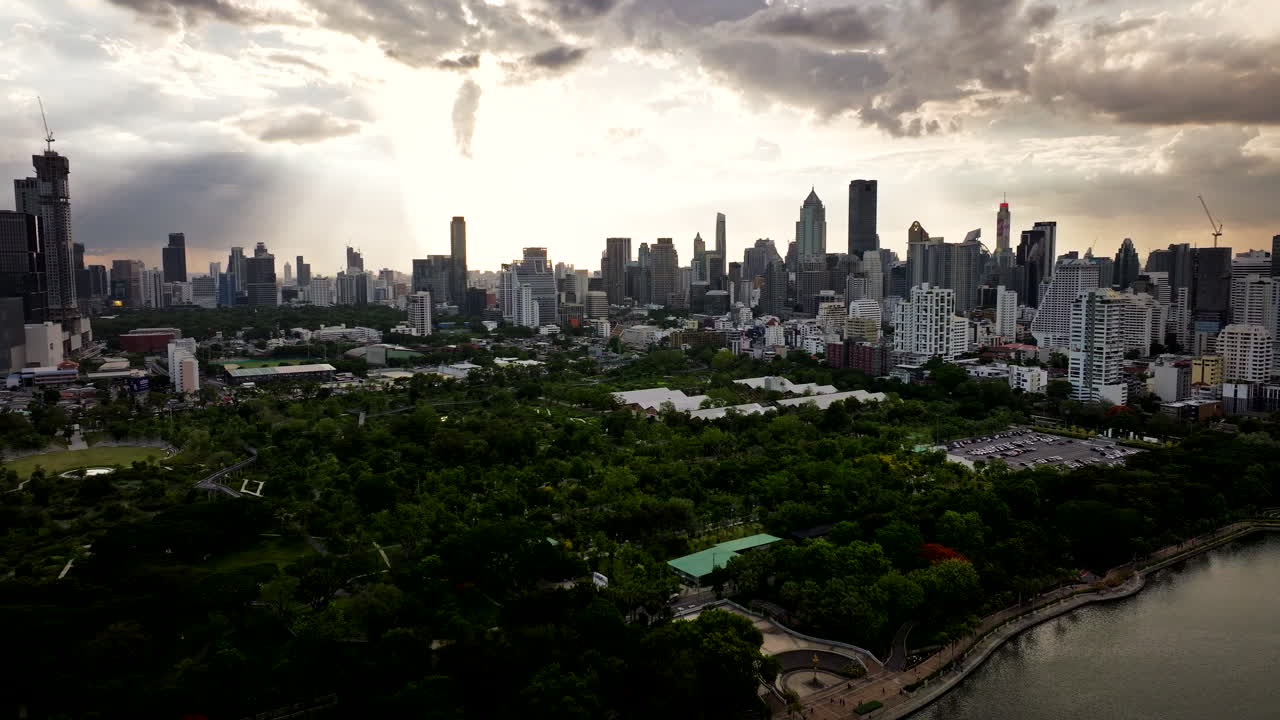 Aerial establishing pullback above Benchakitti Park, lush greenery around pond with Bangkok skyline and sun rays