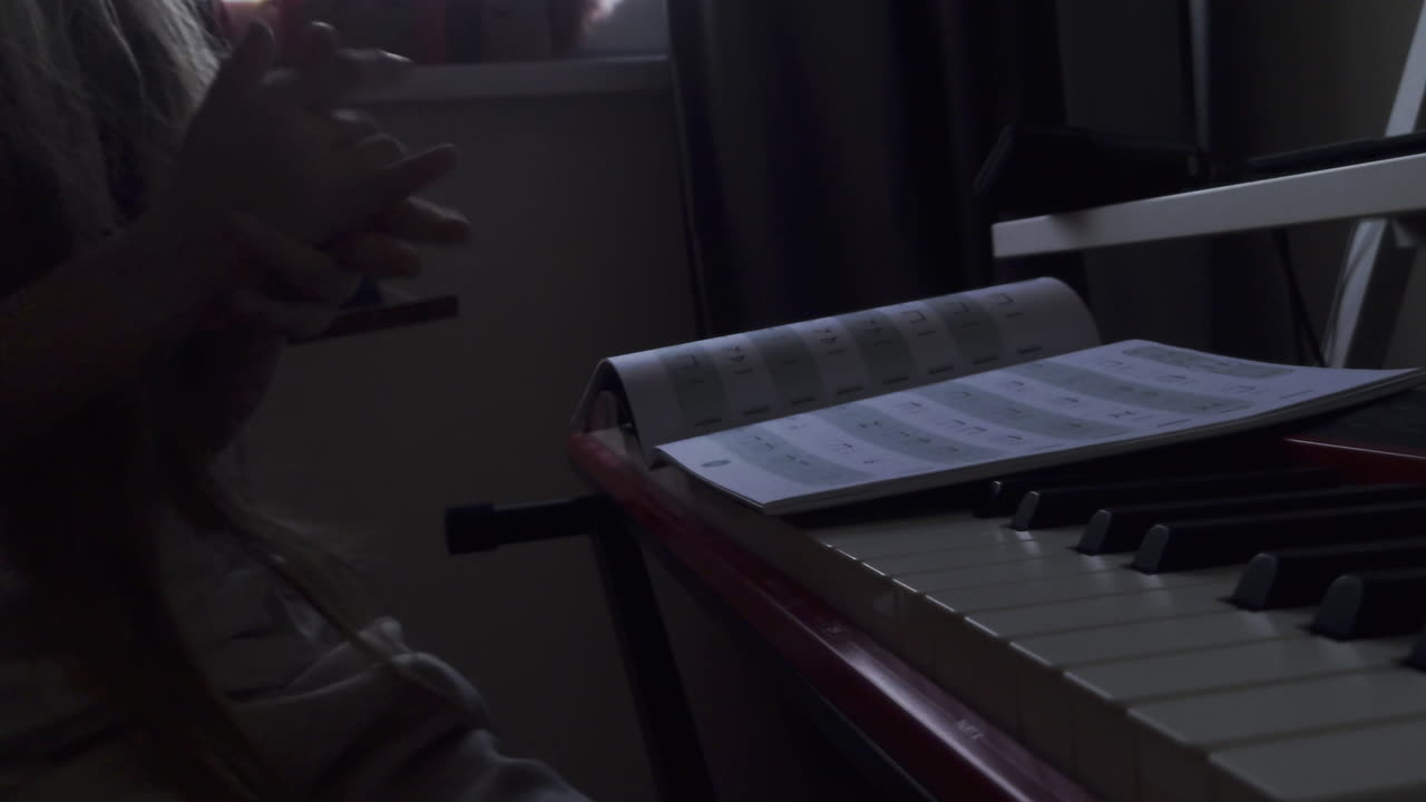 A low-light, static shot capturing a young girl's hands clapping in rhythm as she sits at a digital piano. An open music book rests on the keyboard, emphasizing a focused music theory or rhythm practi
