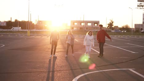 Un-Grupo-De-Cuatro-Amigos-Con-Estilo-Corren-Por-Una-Zona-De-Estacionamiento-Vacía-Al-Aire-Libre-Felicidad-Divertida,-Hombres-Y-Mujeres-Jóvenes-Corren-Al-Atardecer-Y-Luego-Saltan-Juntos-Libremente