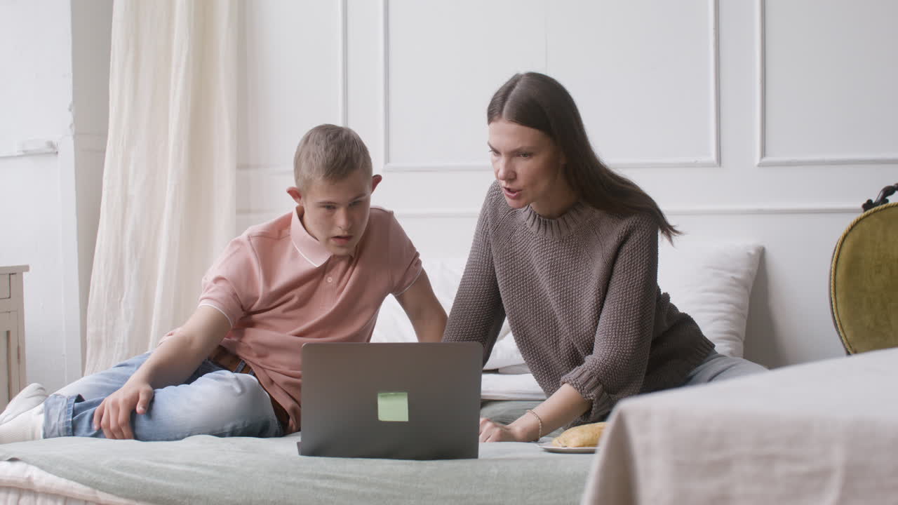 мальчик с синдромом дауна и его мать смотрят что-то на ноутбуке, сидя на кровати в спальне дома