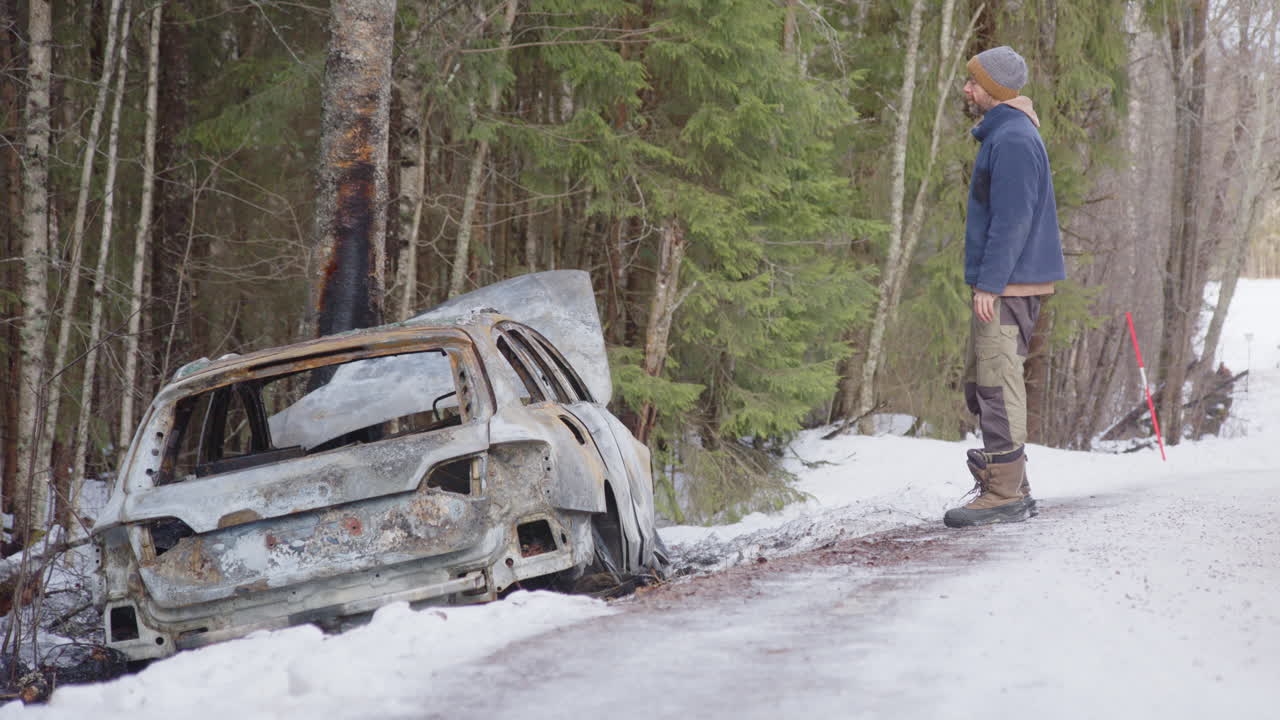 Dejected man stands next to his burned out wreck of a car in snow-covered woods