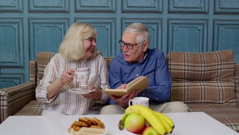 Smiling-senior-couple-grandfather,-grandmother-resting-on-sofa-drinking-coffee,-reading-book-at-home