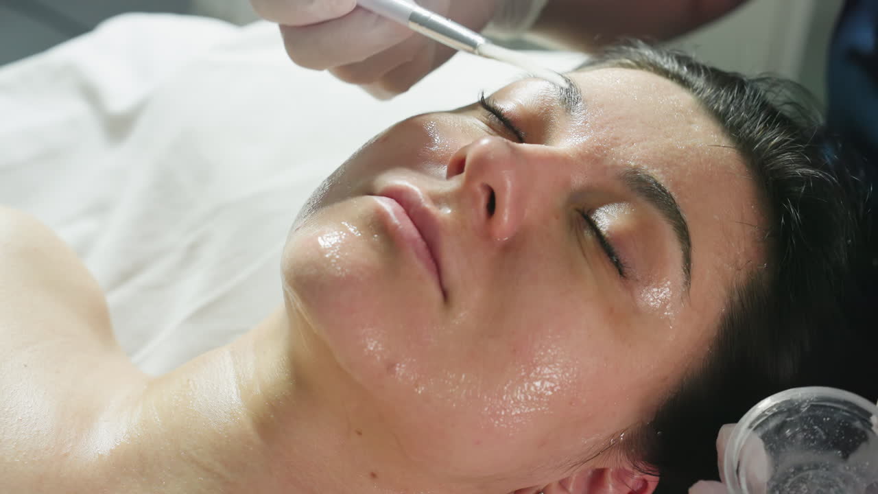 Female client lies with eyes closed on spa bed while beauty expert in gloves dips silicone brush into clear gel and applies smooth nourishing layer across cheek for deep hydration facial treatment