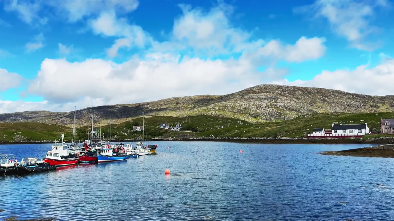 Colourful fishing boats and small vessels moored in the sheltered waters of Scalpay Harbour in Scotland's Outer Hebrides. Rugged hills rise behind the waterfront settlement beneath a bright sky