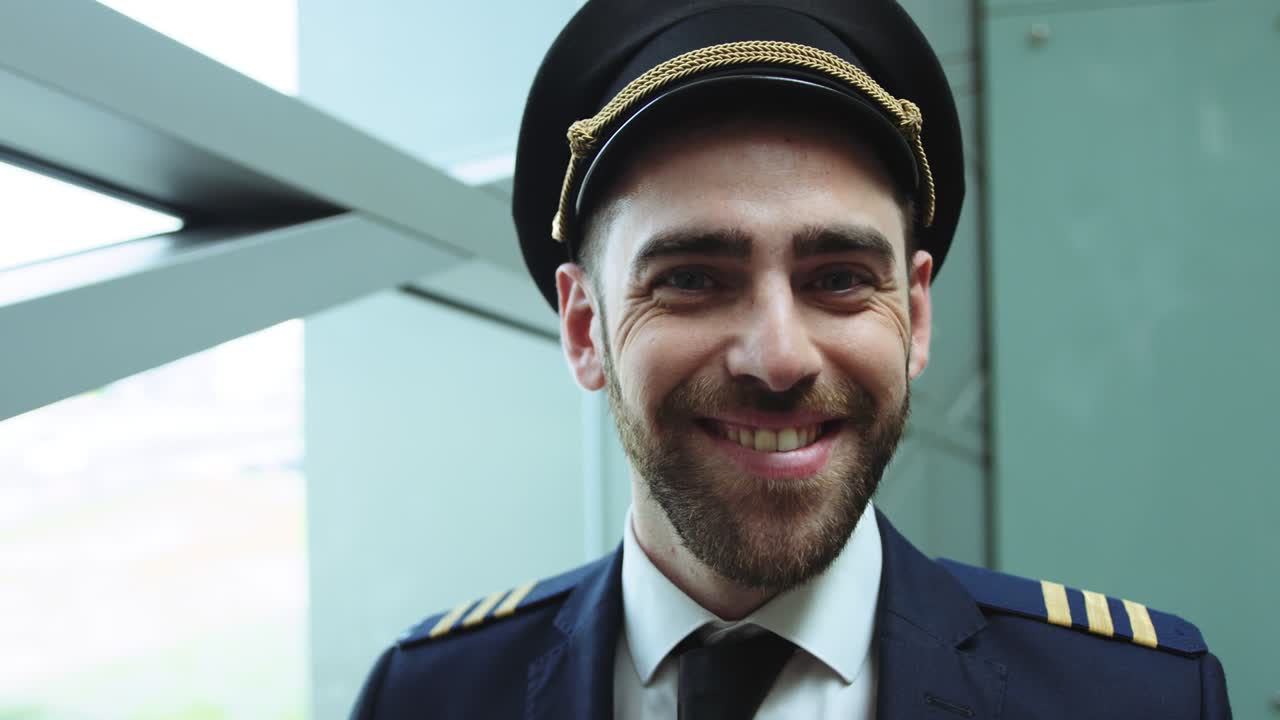 Close up view of strict blue-eyed bearded man in pilot uniform in the hall of airport. Put on cap on head and smiling. Cinematic shot on RED camera.