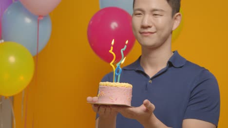 Studio-Portrait-Of-Man-Wearing-Party-Hat-Celebrating-Birthday-Holding-Cake-With-Candles