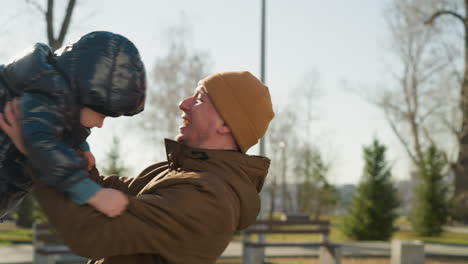 a close-up of a father and son enjoying outdoor playtime as the father spins his son gently in a park and placed him on his shoulders, with trees and benches in the background