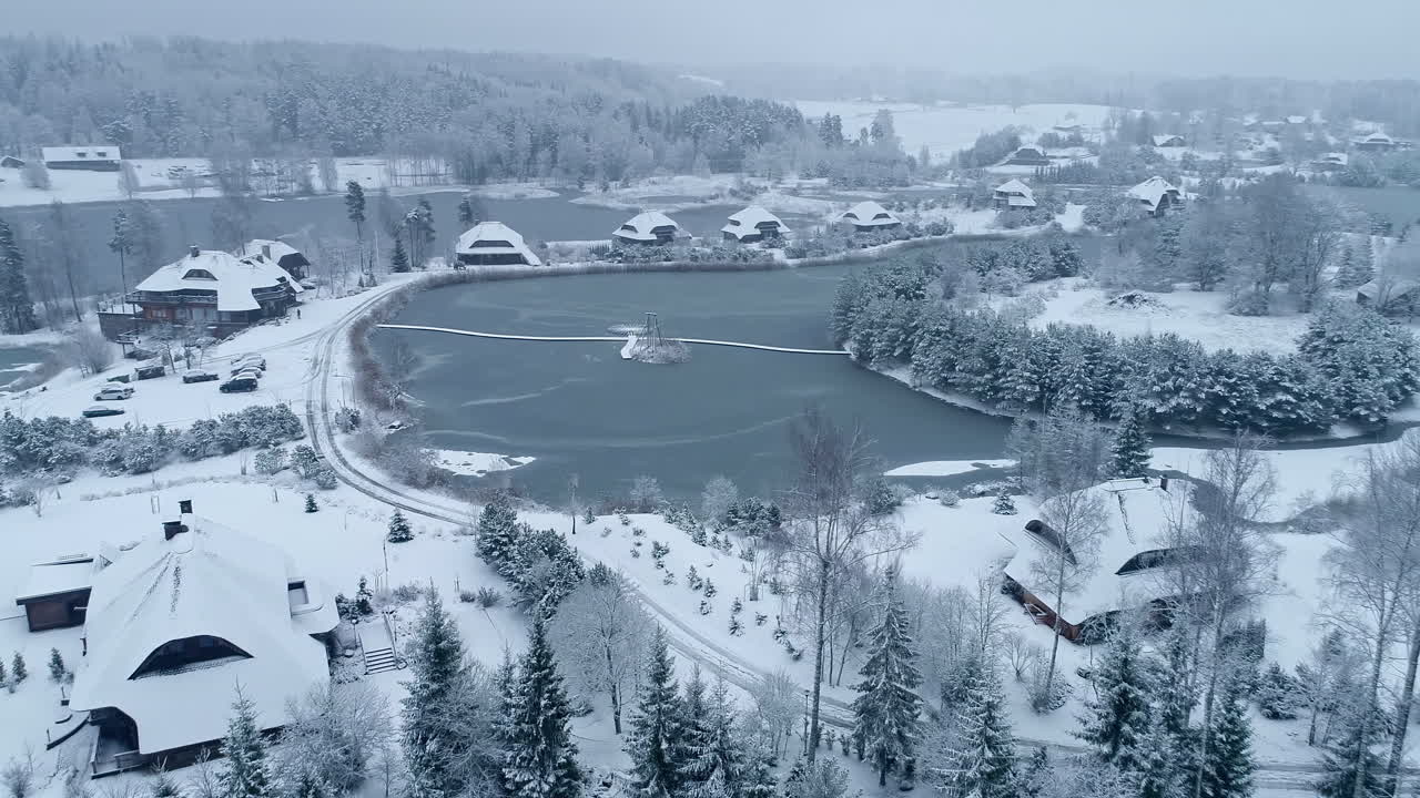 추운 겨울날 얼어붙은 호수 옆에 눈으로 덮인 나무 오두막 위로 공중 드론이 앞으로 이동합니다.