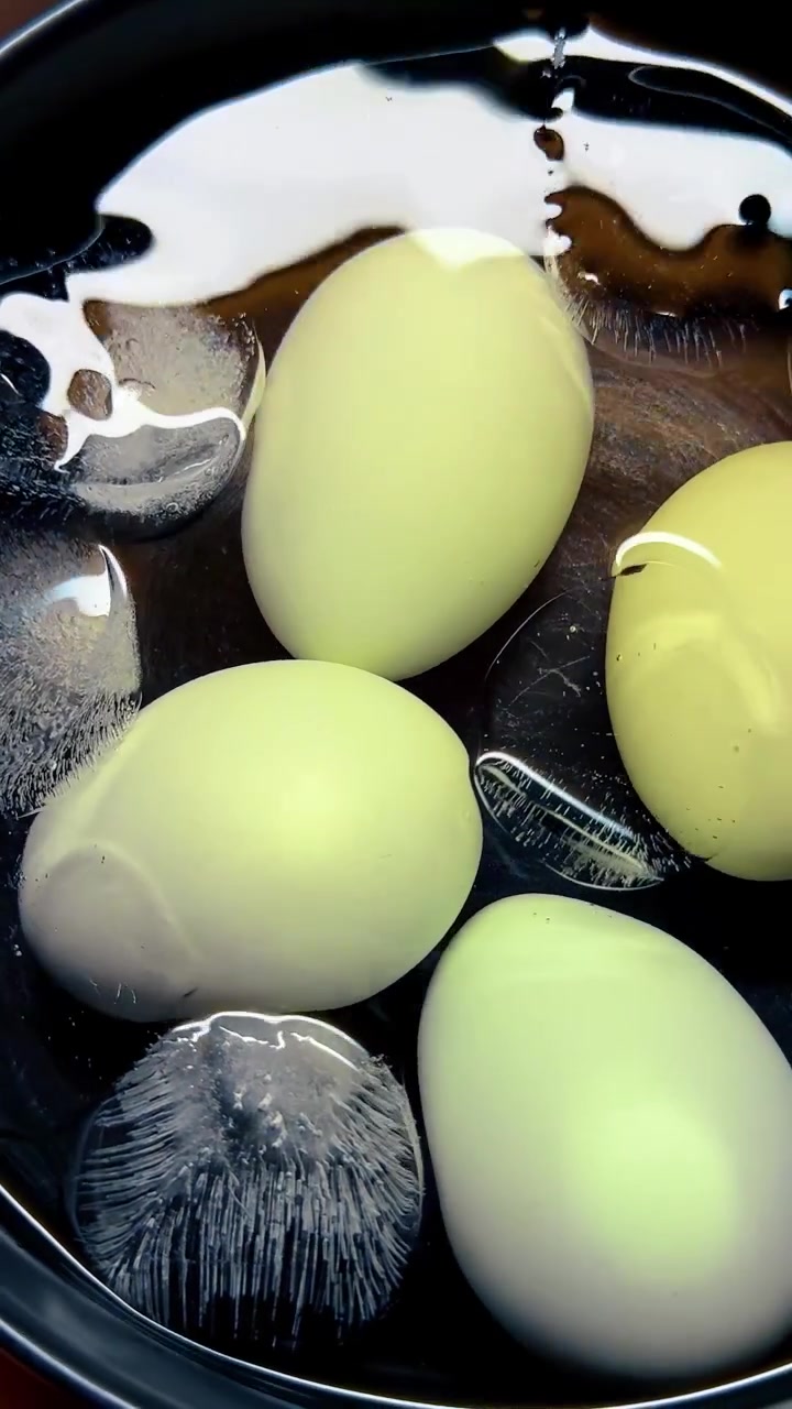 Light blue boiled eggs cooling in bowl ice water. Top view of light blue eggs submerged in cold water with ice cubes in black bowl.