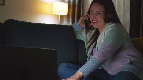 smiling woman sitting on sofa at home at night talking on mobile phone and watching movie or show on laptop 9