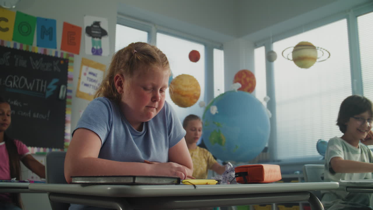 Stressed Primary School Girl Sitting at Desk Looking at Camera While Aggressive Classmates Abusing