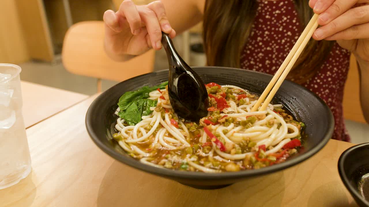 Young woman stirs spicy noodle soup in black bowl, warm restaurant lighting, close-up shot