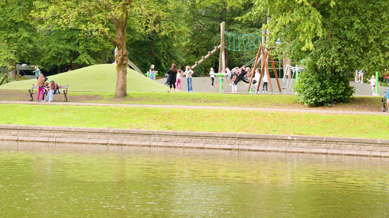Children and adults interact at a lively playground by a calm lake in bright daylight