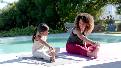 happy biracial mother and daughter practicing yoga sitting stretching in sunny garden, slow motion