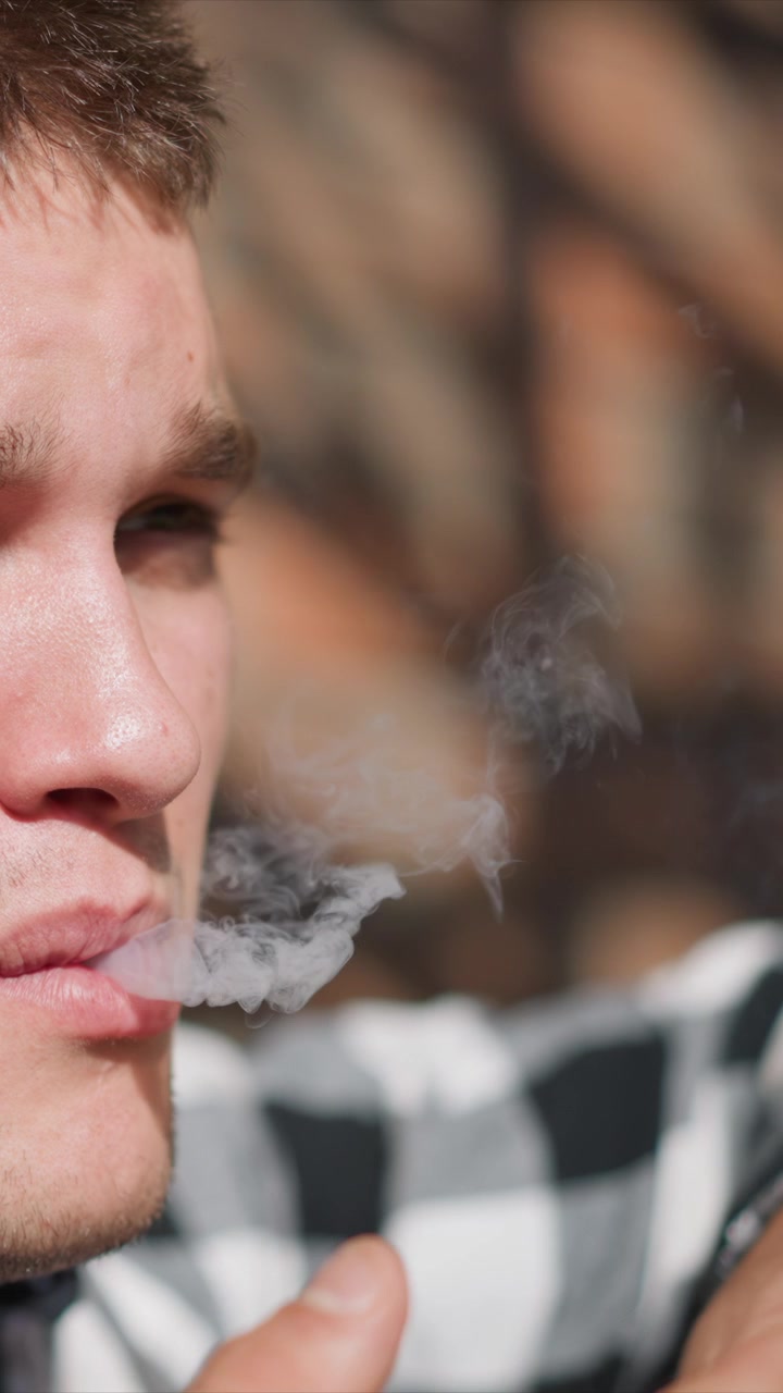 Primer plano de un hombre fumando, sosteniendo y llevando un cigarrillo a su boca. El sujeto lleva una camisa a cuadros con detalles de textura visibles como poros de la piel o pecas, con iluminación suave.