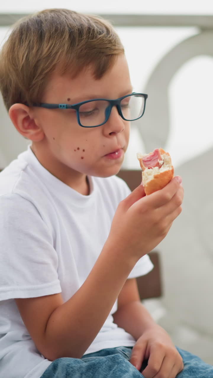A little boy with glasses is captured eating his snack with a mouth full, enjoying every bite, a blurred view of another child also with a full mouth with his snacks