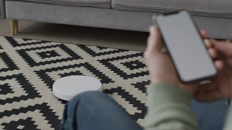 Unrecognizable Old Man Sitting On Sofa And Using Mobile Phone To Control A Robot Vacuum Cleaner