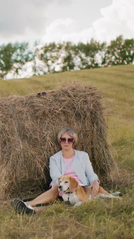 mujer con gafas de sol y traje casual se sienta en tierras de cultivo cubiertas de hierba, apoyándose en la bala de heno mientras acaricia suavemente al perro, campo abierto con colinas onduladas, cielo nublado y entorno rural pacífico