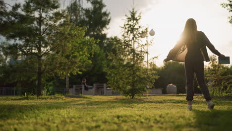 lady joyfully turns outdoors in grassy field with book in hand, as sunlight creates a radiant atmosphere around her, background features greenery, trees, and a slightly blurred view of someone seated