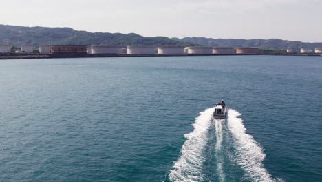 Aerial-drone-follows-motorboat-from-below-sailing-blue-Japanese-Sea,-coastline-with-mountain-range-background,-Japan-Travel-destination-Wakayama